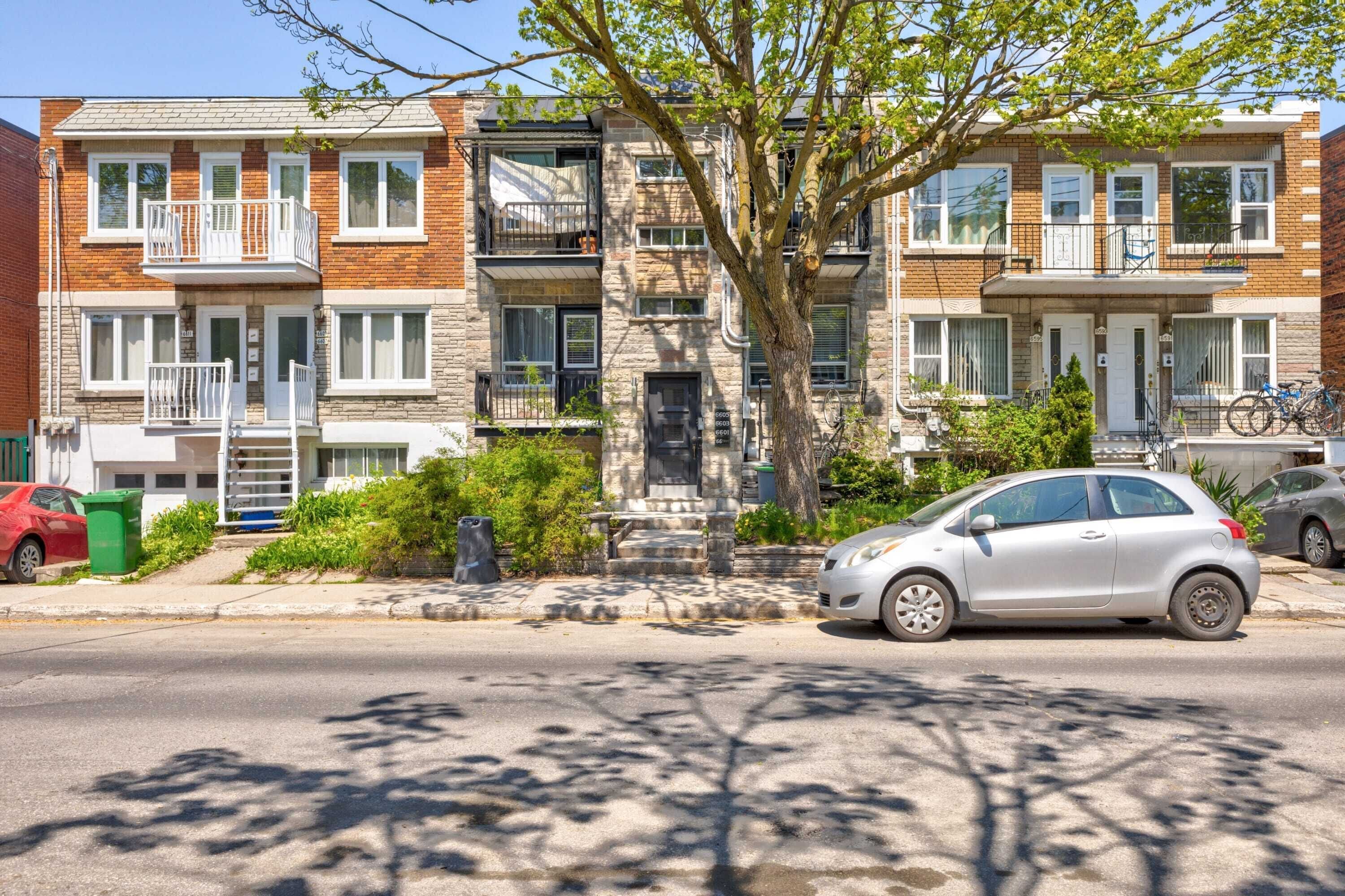 Une rangée de maisons de ville avec des porches à l’avant, un arbre au centre et une voiture argentée garée dans la rue.