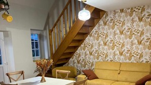 Un comedor con una mesa blanca, sillas de madera y un florero con flores secas. Se ve un sofá amarillo y una escalera de madera al fondo.