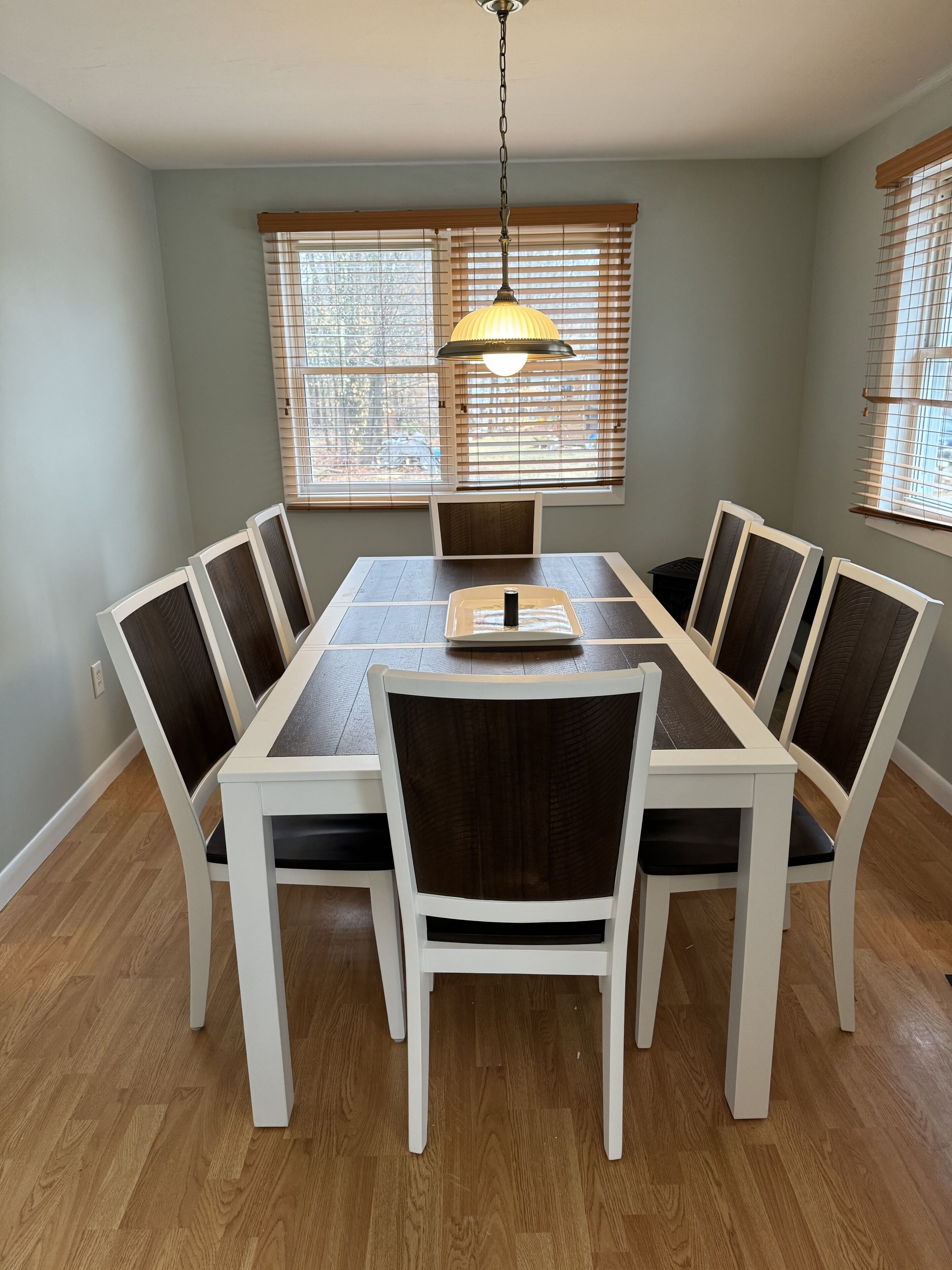 A dining room with a rectangular table surrounded by six chairs, a hanging light fixture, and two windows with blinds.