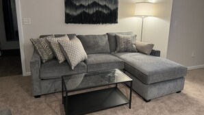 A grey sectional sofa with patterned cushions, a glass coffee table, a wall-mounted tapestry, and a floor lamp.