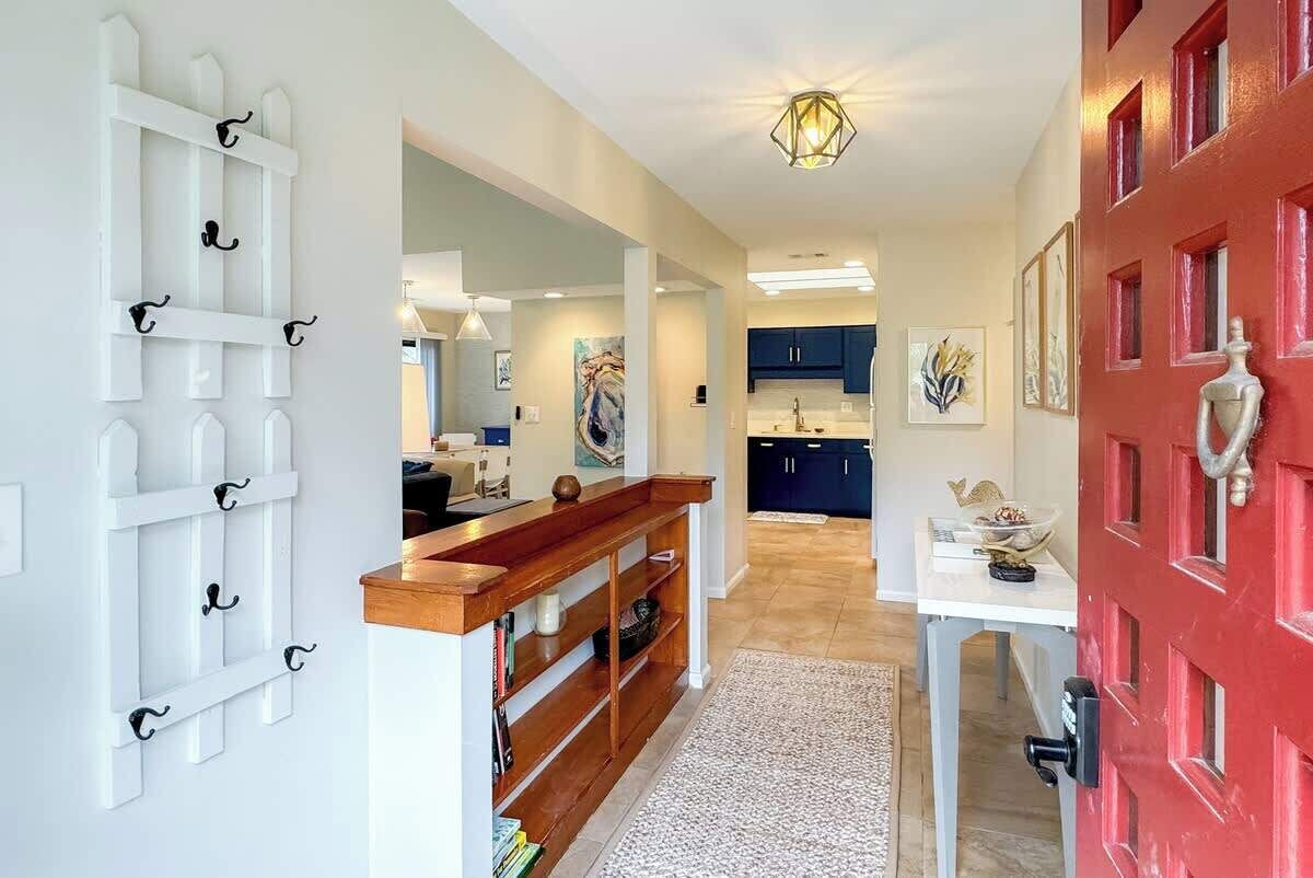 A hallway with a white picket fence, a wooden bench, a red door, and a kitchen area in the background.