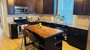 A modern kitchen with dark wood cabinets, stainless steel appliances, a wooden island with bar stools, and a window with blinds.