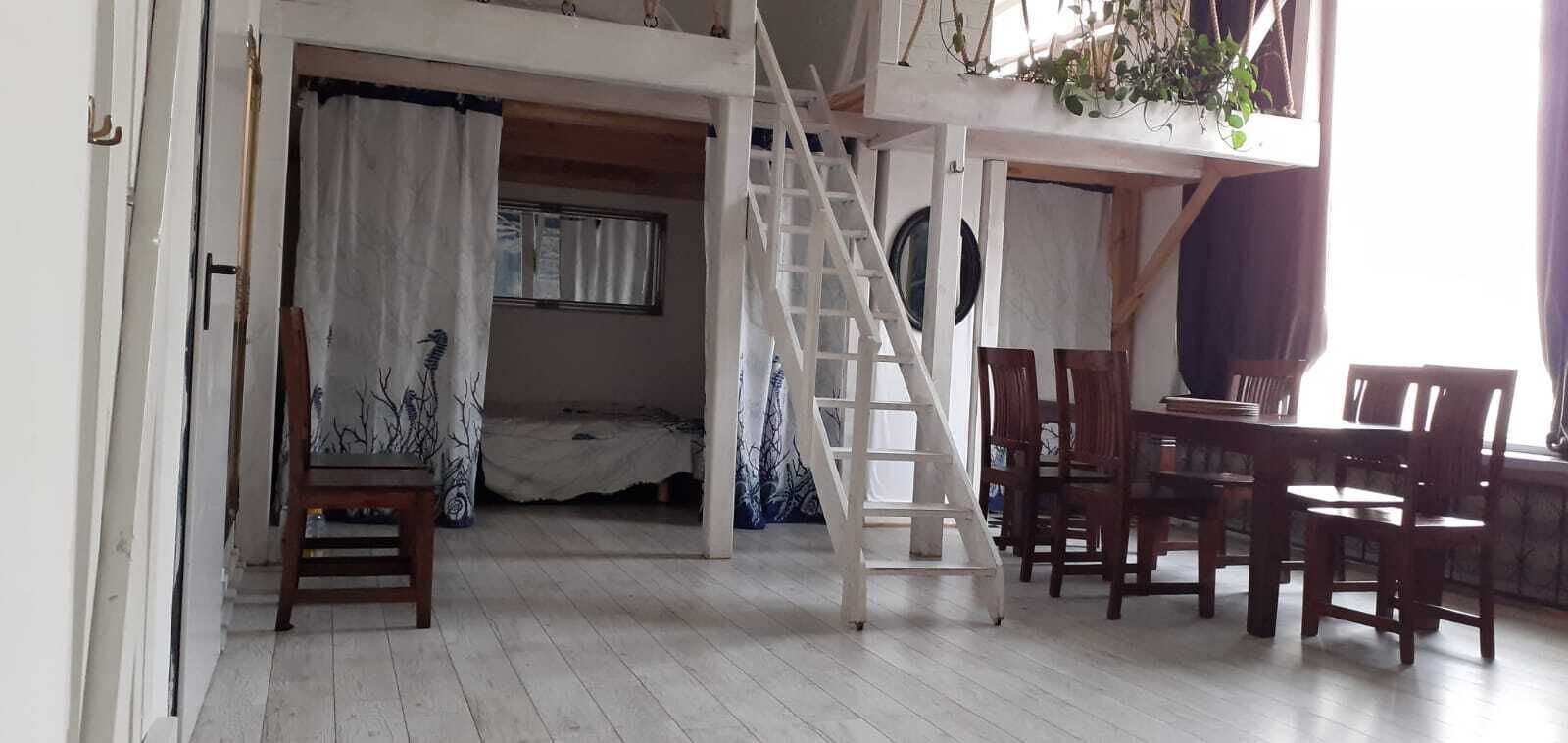 A room with a wooden floor, a bed with a white and blue patterned bedspread, a wooden chair, a dining table with chairs, and a white ladder leading to a loft area with a window and a hanging plant.