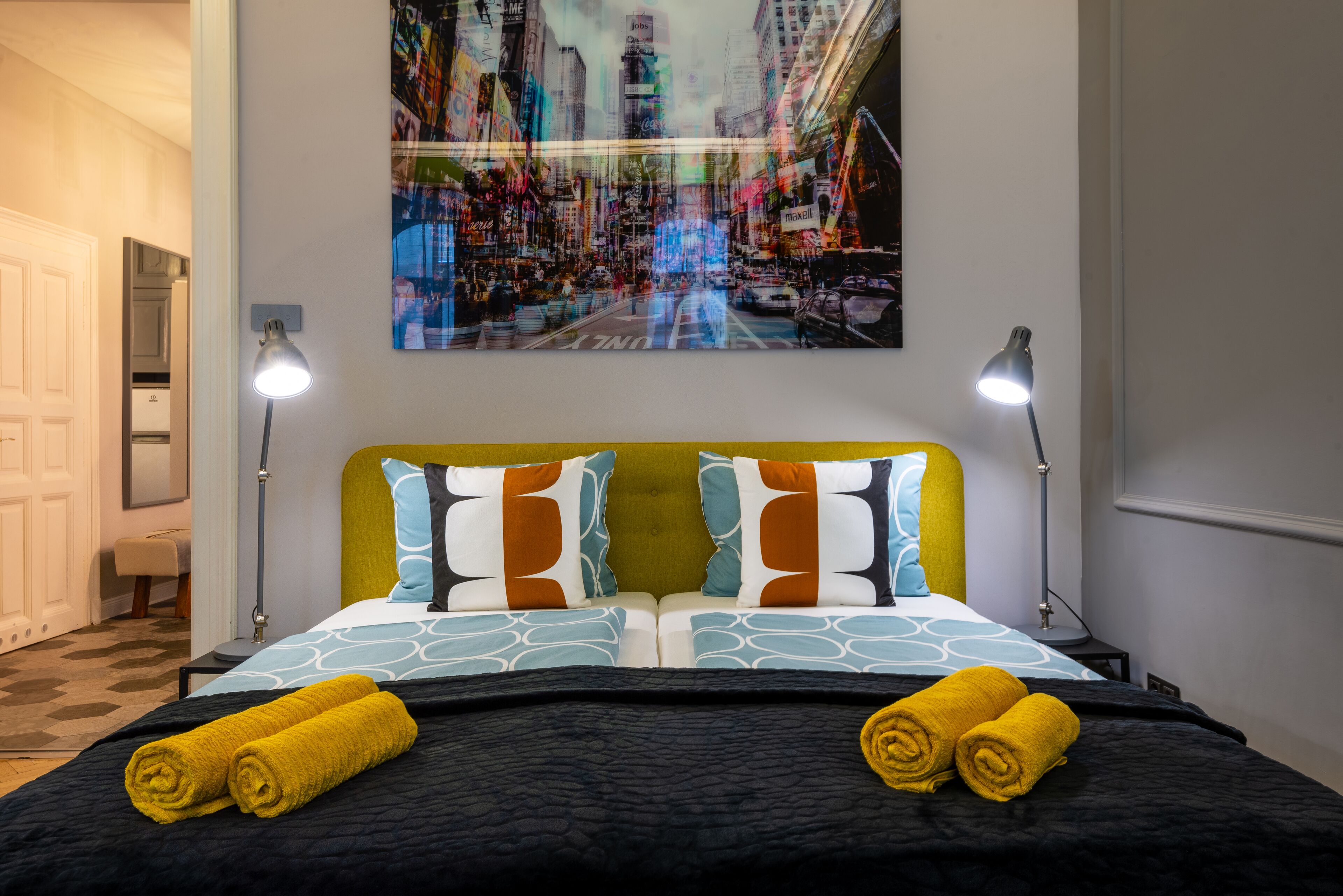 Ett hotellrum med en säng, två sänglampor, ett konstverk med en stadsbild på väggen och två hoprullade handdukar på sängen.