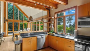 A modern kitchen with wooden cabinets, a dishwasher, and a large window overlooking a forested area. - Curated art, luxe linens & indoor-outdoor fireplaces. (Stevenson)