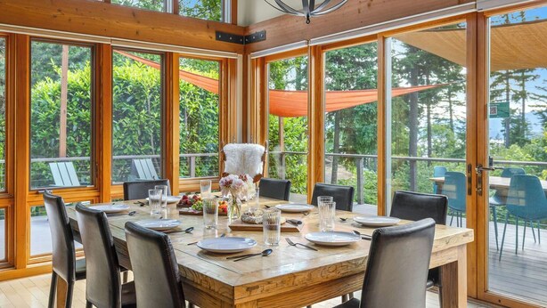A dining area with a wooden table set for six, surrounded by large windows offering a view of greenery. - Curated art, luxe linens & indoor-outdoor fireplaces. (Stevenson)