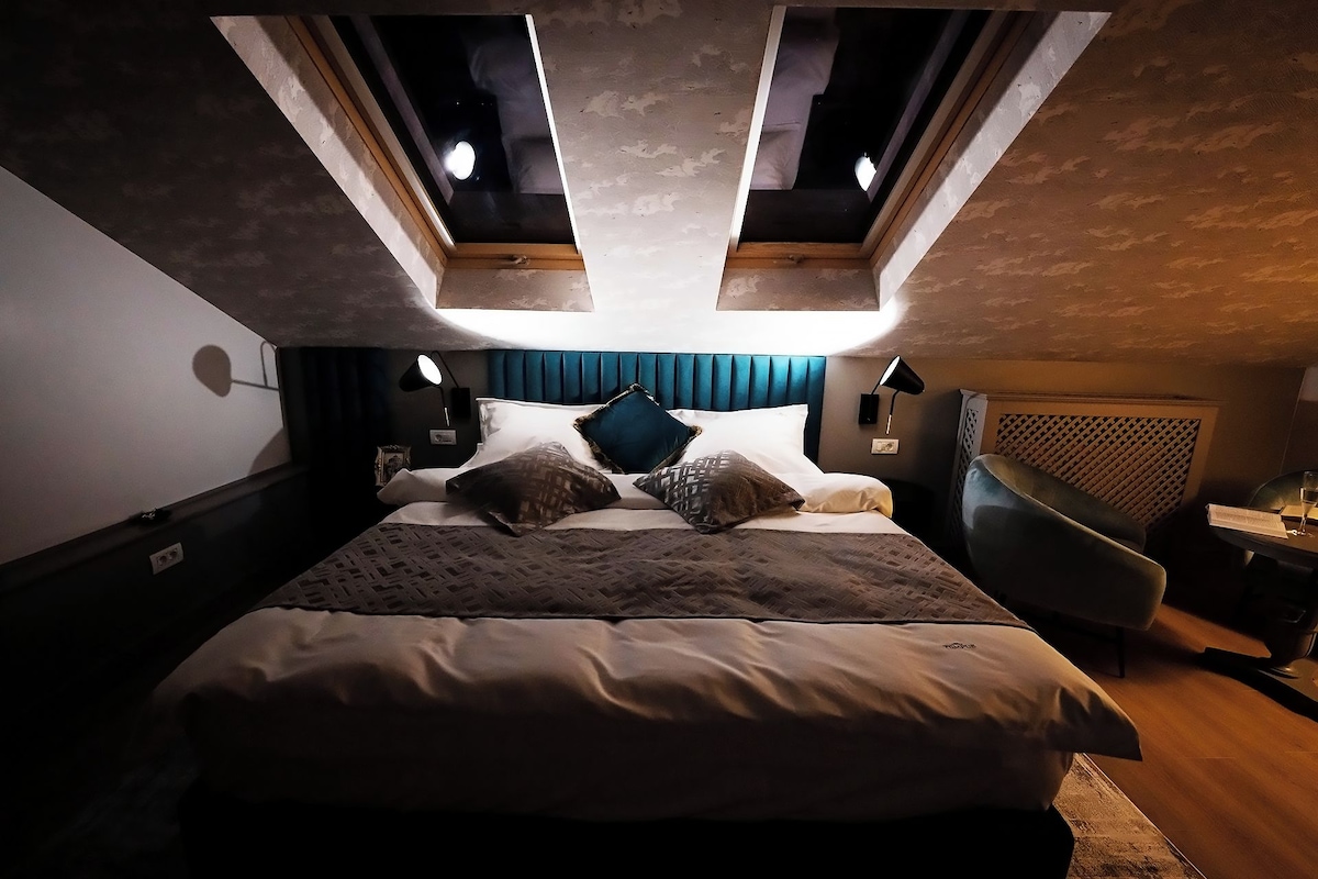 Une chambre à coucher avec un lit, des oreillers, une table de chevet, une lampe, une chaise et une fenêtre donnant sur le ciel.
