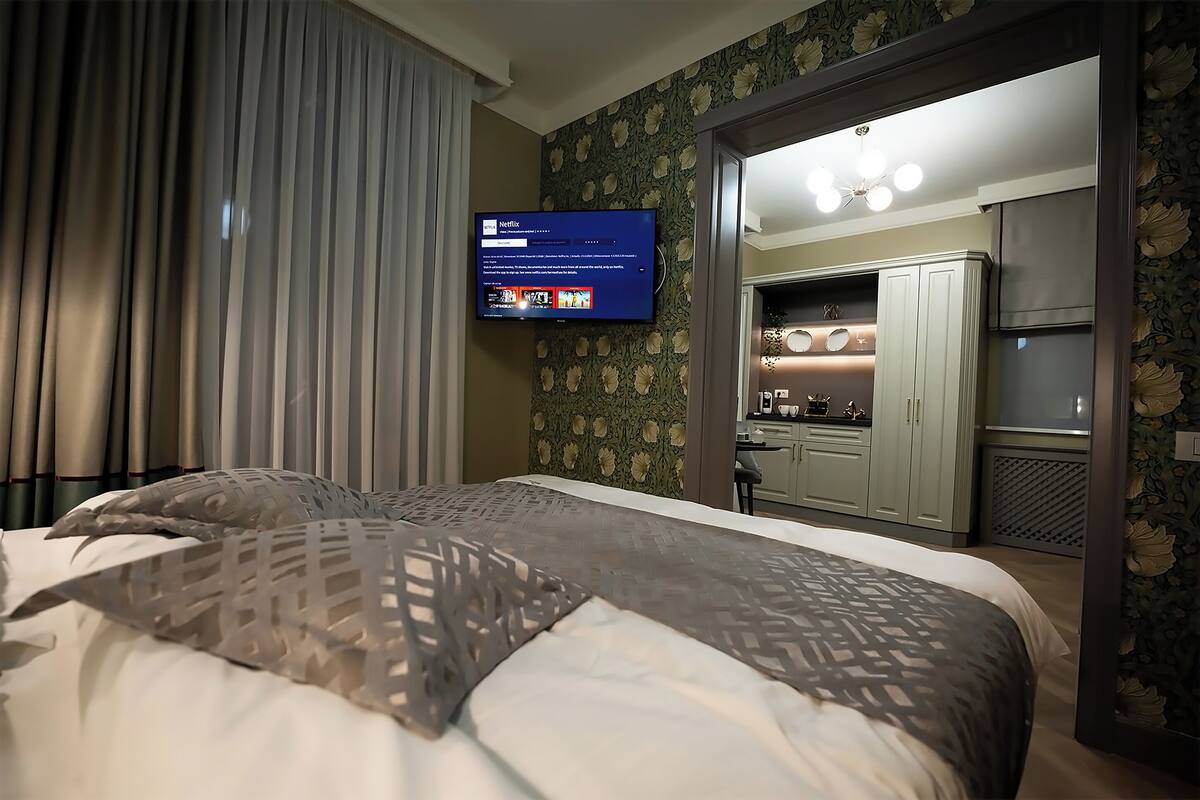 A hotel room with a bed, curtains, a TV on the wall, and a kitchenette area.