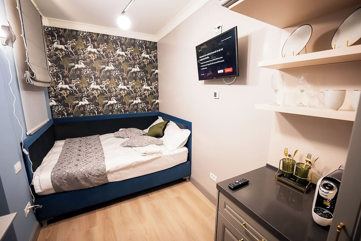 A bedroom with a blue bed, a television on the wall, and a shelf with cups and a remote control.