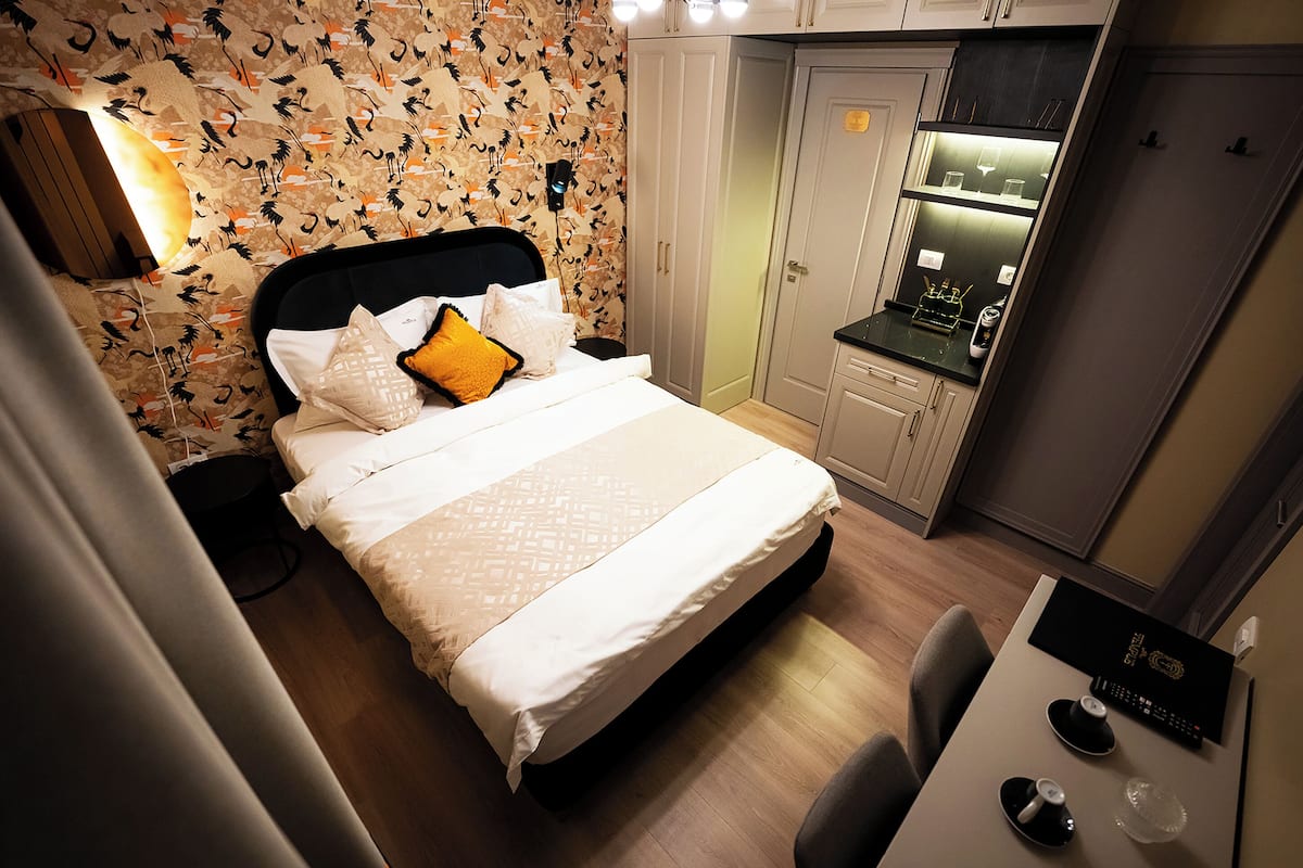 A neatly made bed with a patterned bedspread, a bedside table with a remote control, and a small desk with a cup and saucer.