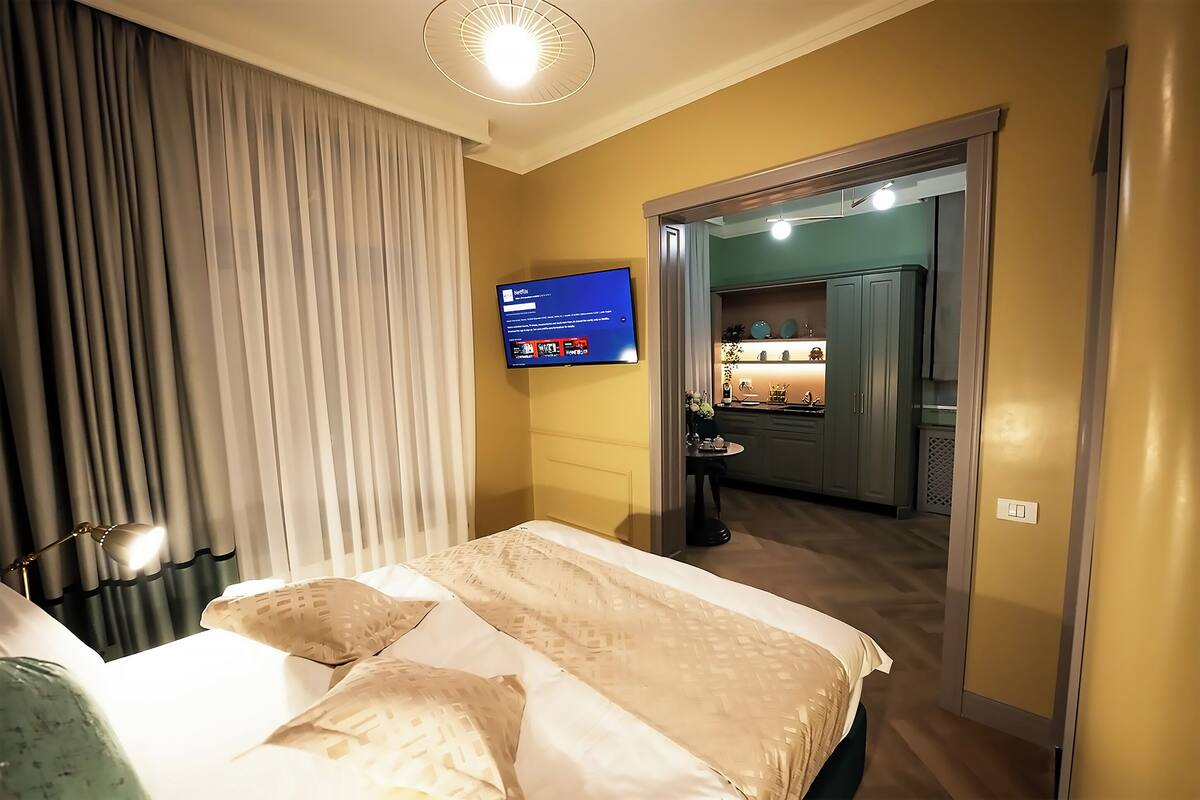 A hotel room with a bed, a TV mounted on the wall, and a view into the kitchen area.