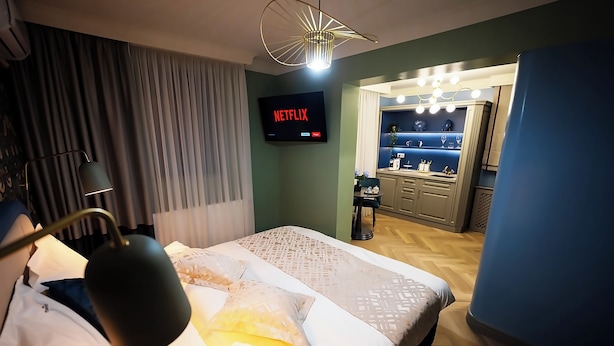 Quarto de hotel com cama, TV exibindo 'Netflix', escrivaninha e toucador com iluminação.
