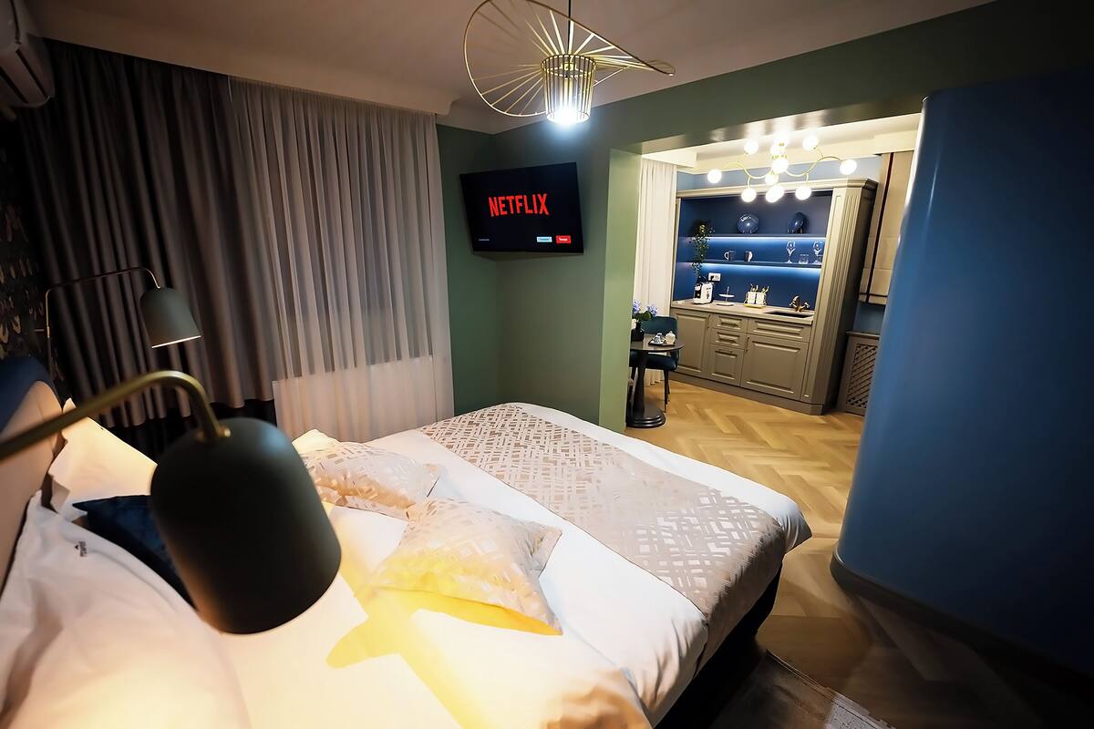 A hotel room with a bed, a TV displaying 'Netflix', a desk, and a vanity with lights.