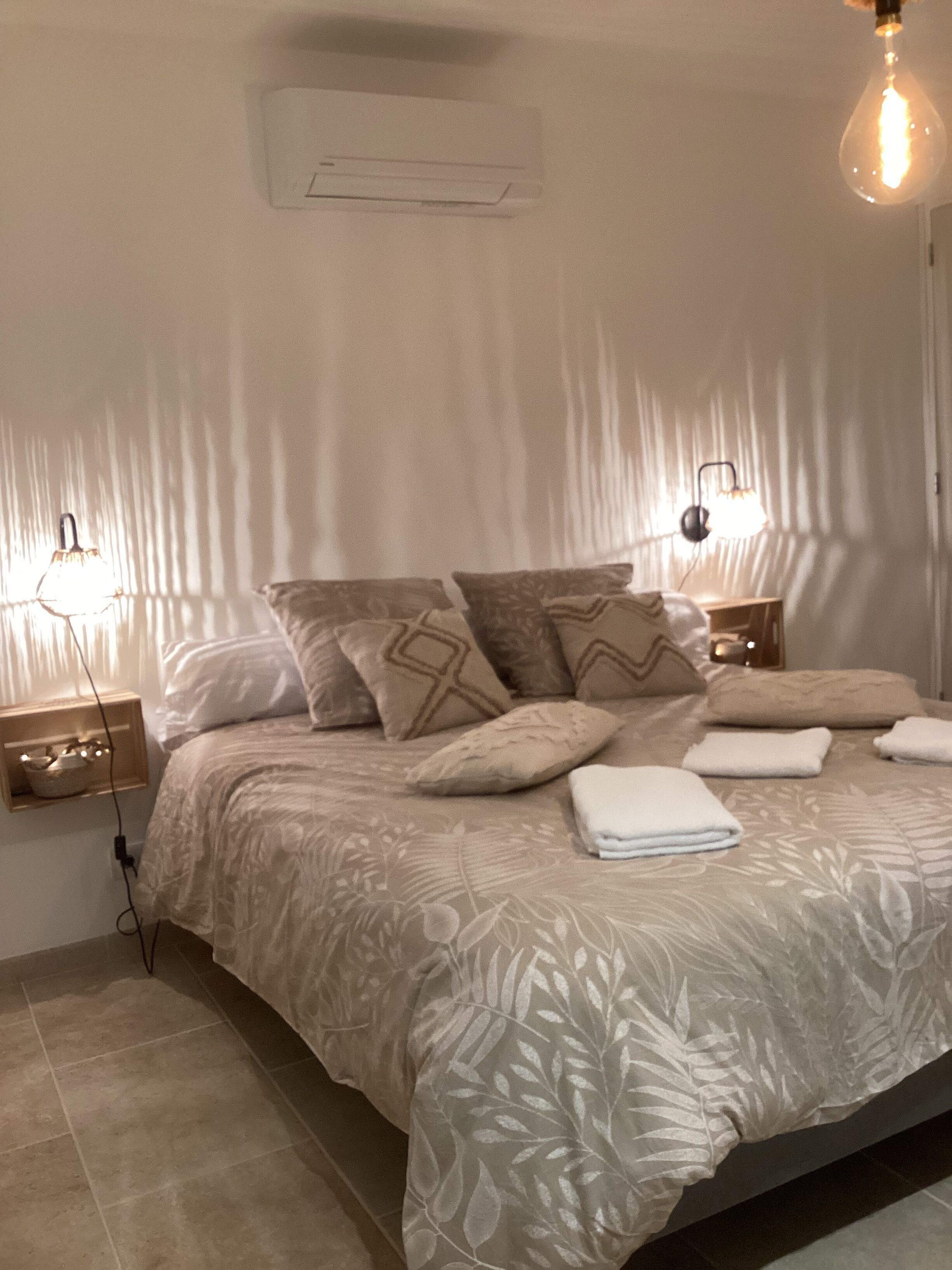 Un dormitorio con cama, almohadas, mesita de noche, lámpara y aire acondicionado instalado en la pared.