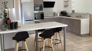 A modern kitchen with a white countertop, stainless steel appliances, and two black bar stools. - Gîte rdc Villa - Village Provençal - Proche Uzès (Le Pin)