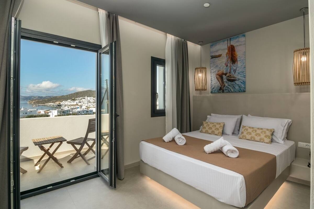 A hotel room with a bed, a view of a coastal town, and a balcony with chairs.