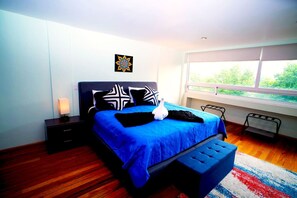Une chambre moderne avec un grand lit, une table de chevet, une commode, une fenêtre donnant sur un paysage verdoyant et un tapis coloré.