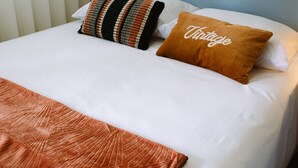 Una cama bien hecha con sábanas blancas, un plaid marrón y dos almohadas decorativas, una con estampado a rayas y la otra con la palabra 'Vintage' bordada.