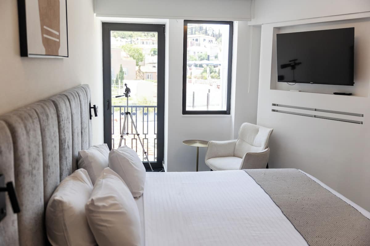 Une chambre d’hôtel moderne dotée d’un grand lit, d’un téléviseur fixé au mur, d’un balcon offrant une vue sur la ville et d’une petite table avec une chaise.