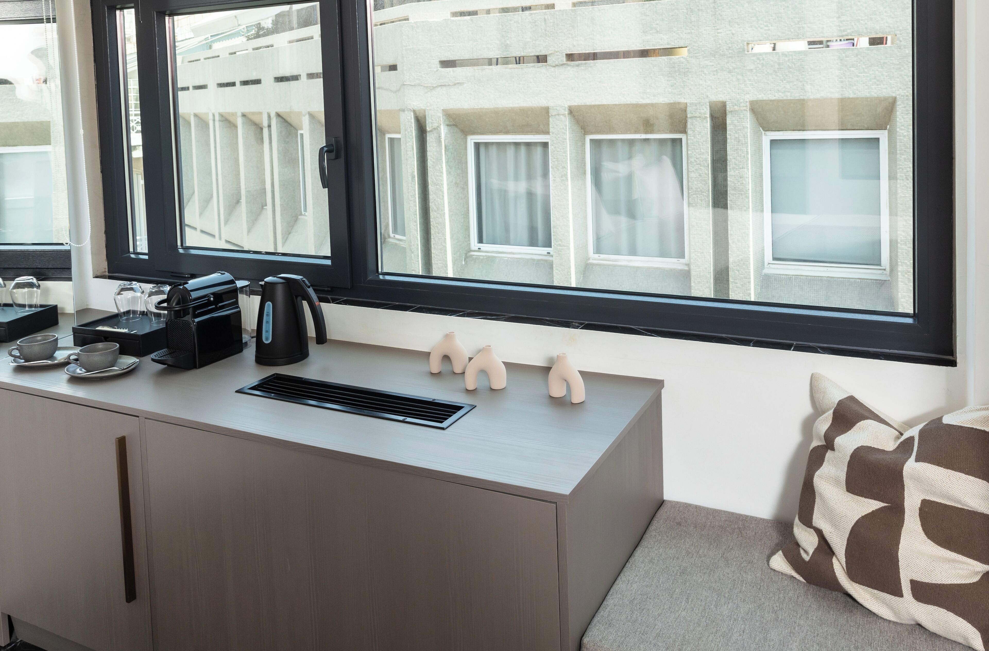 A modern hotel room with a minimalist desk, a coffee maker, and a view of a building through the window.