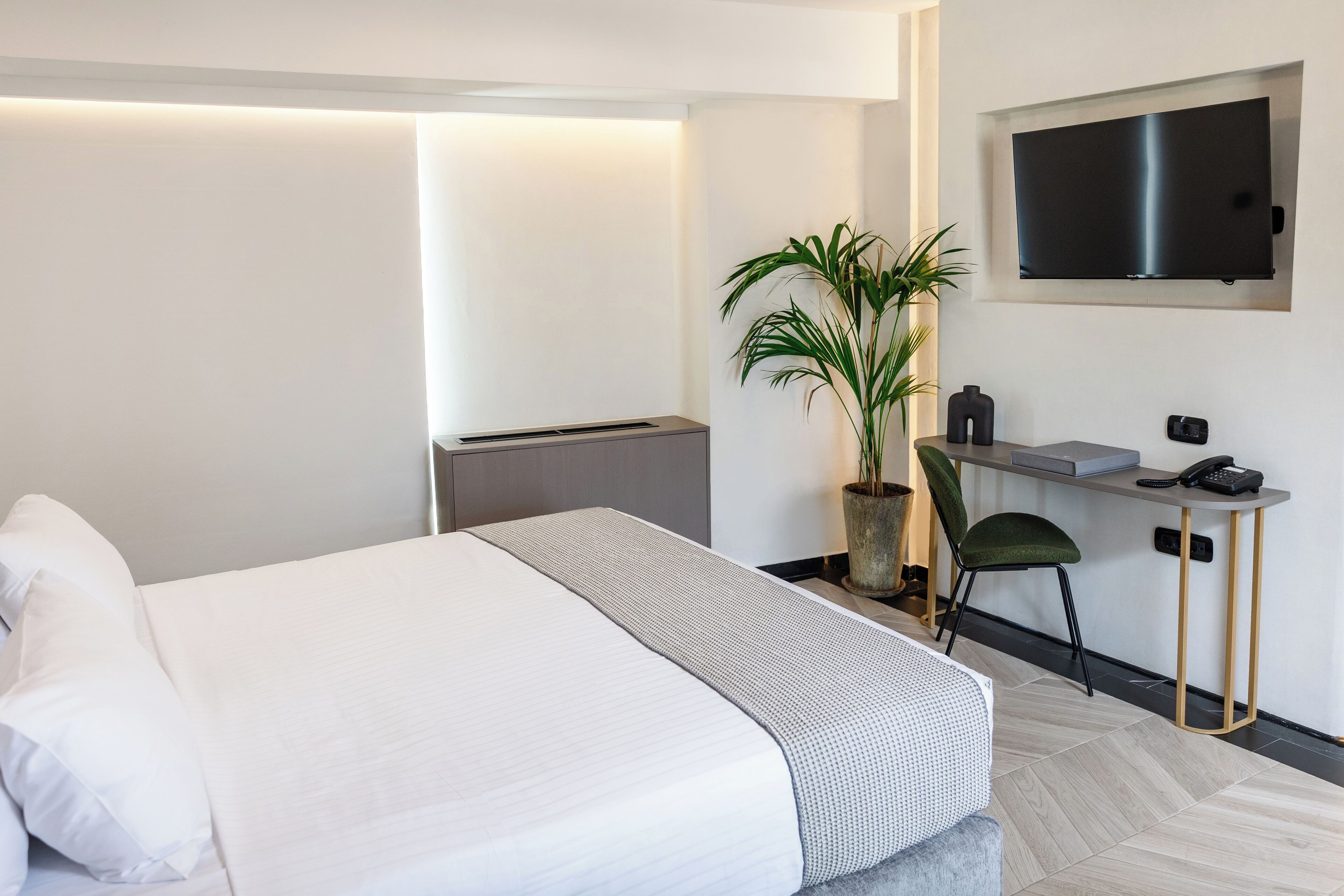 A modern hotel room with a bed, a desk, a chair, a potted plant, and a flat-screen TV mounted on the wall.