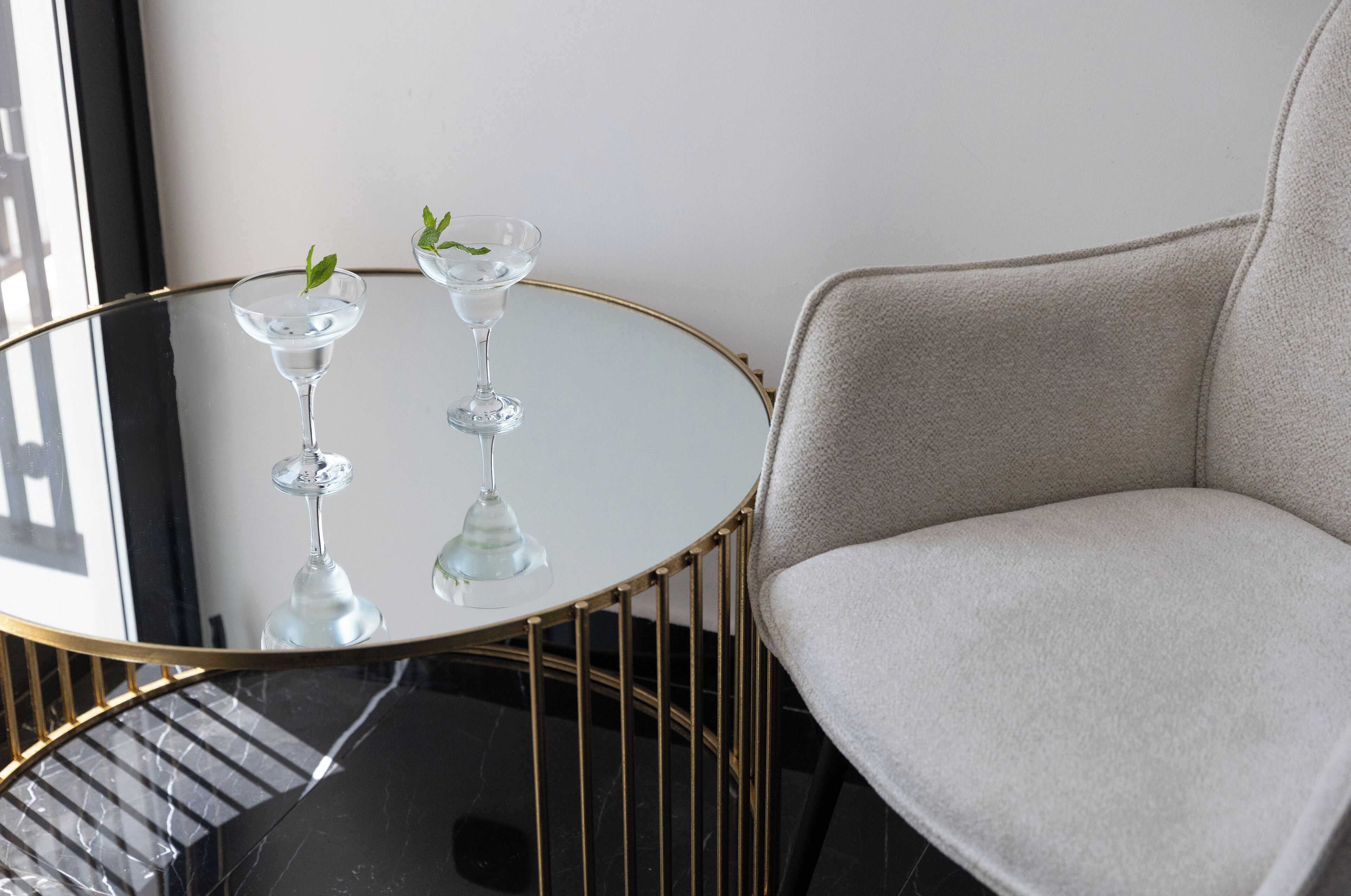 A round glass table with two martini glasses, a gold-framed chair, and a view of a building outside.