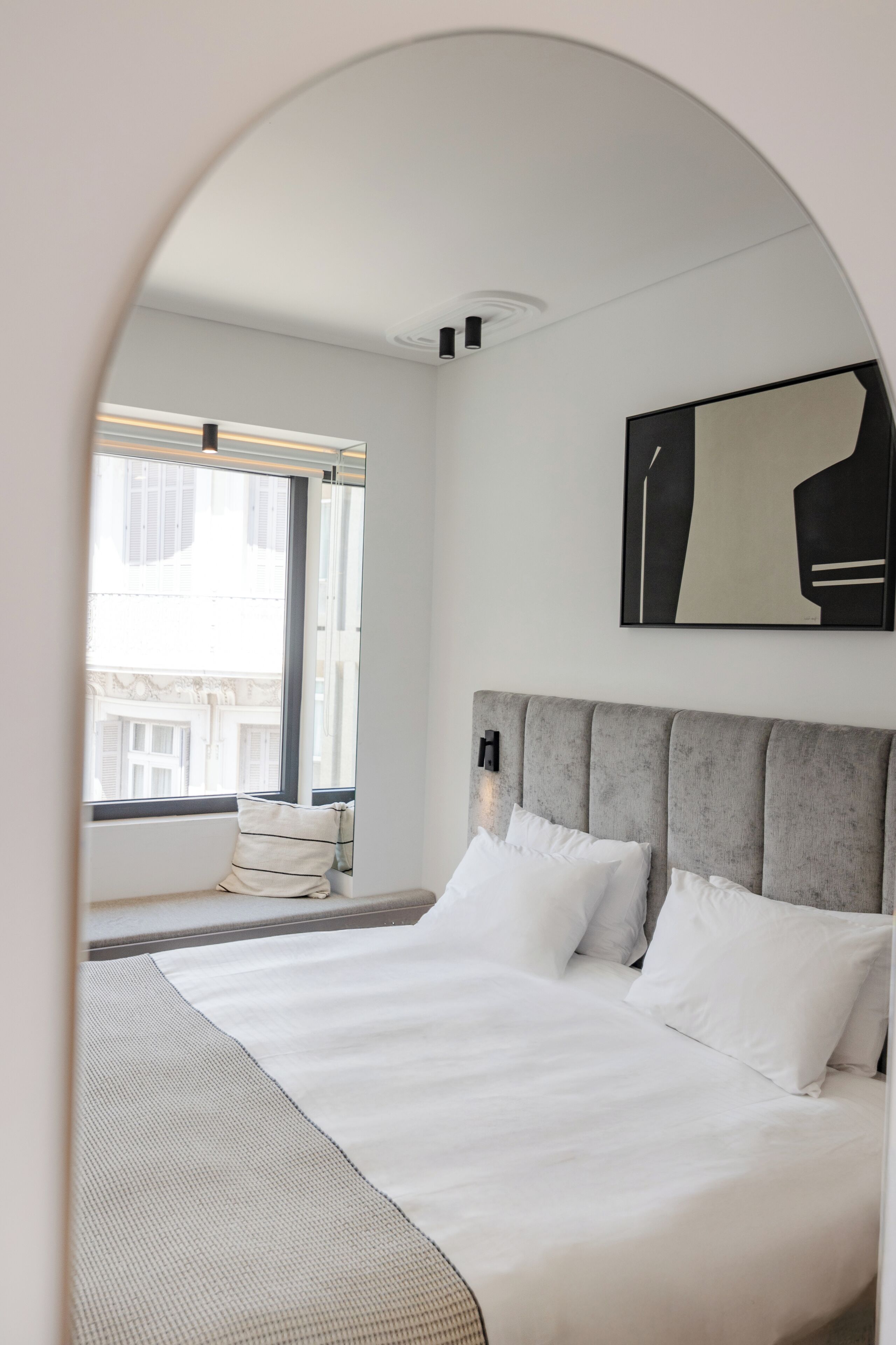 A modern bedroom with a large bed, a window with blinds, a grey upholstered headboard, and a minimalist artwork on the wall.
