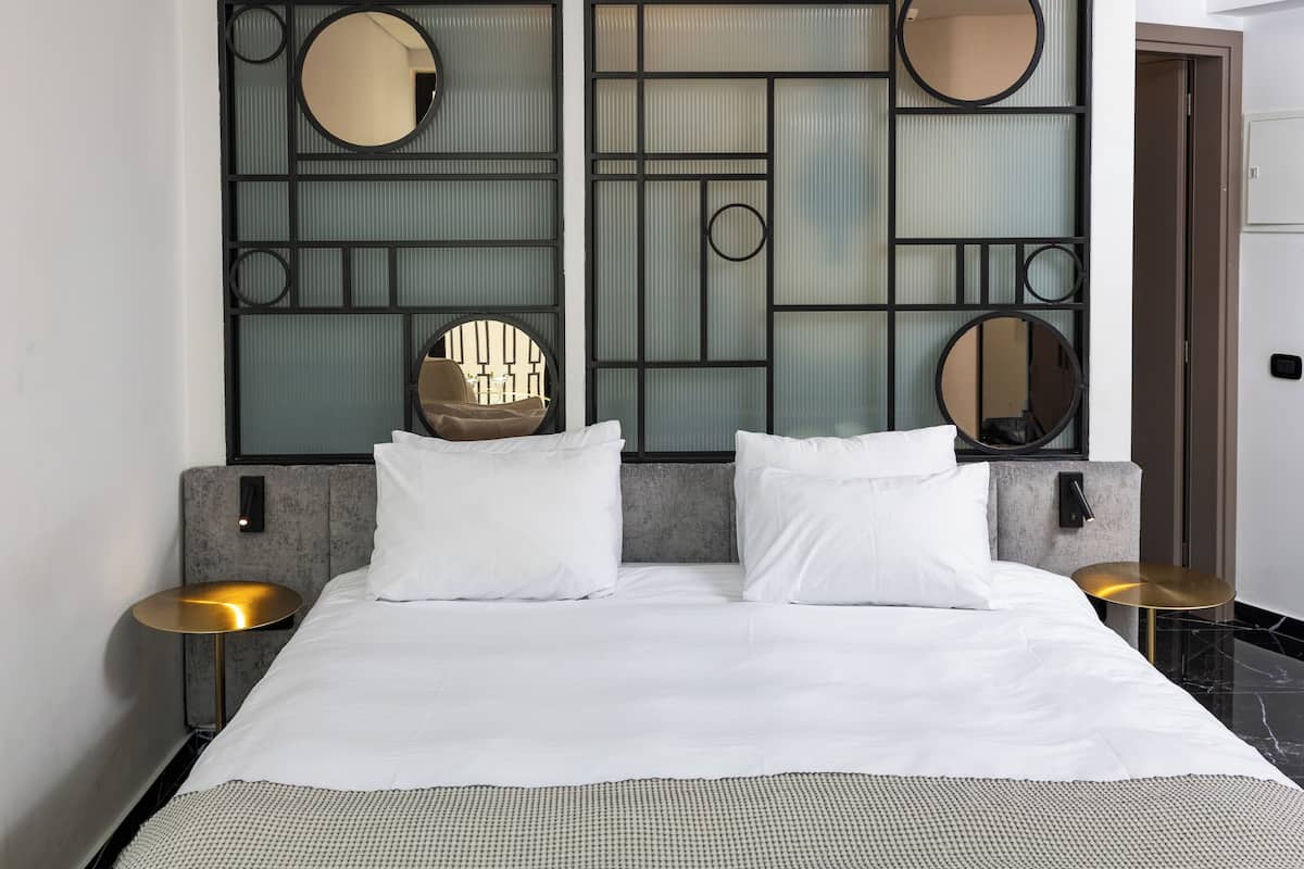 Une chambre d’hôtel moderne, dotée d’un grand lit, de deux tables de chevet et d’un panneau mural décoratif agrémenté de miroirs et de motifs géométriques.