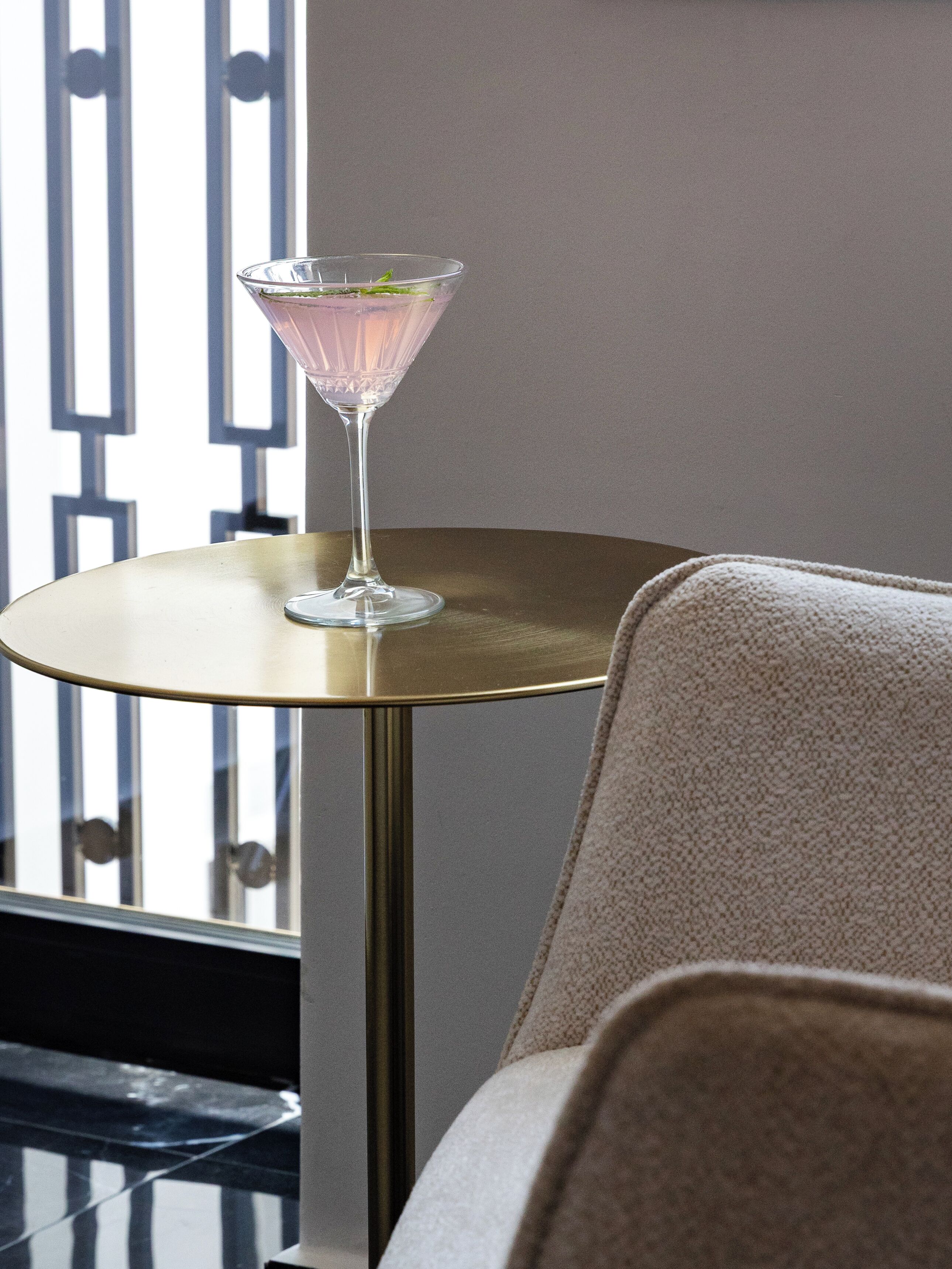 Un verre à martini posé sur une table ronde dont le pied est doré, placé près d’une fenêtre munie de stores verticaux.