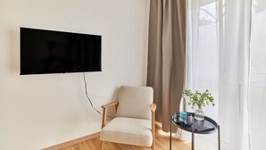 A living room with a flat-screen TV mounted on the wall, a beige armchair, a small round table with a plant, and a window with curtains.