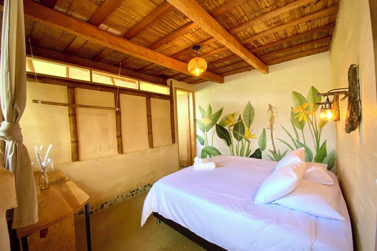 A bedroom with a wooden ceiling, a bed with white bedding, a nightstand with a bottle and glasses, and a wall mural of tropical plants.