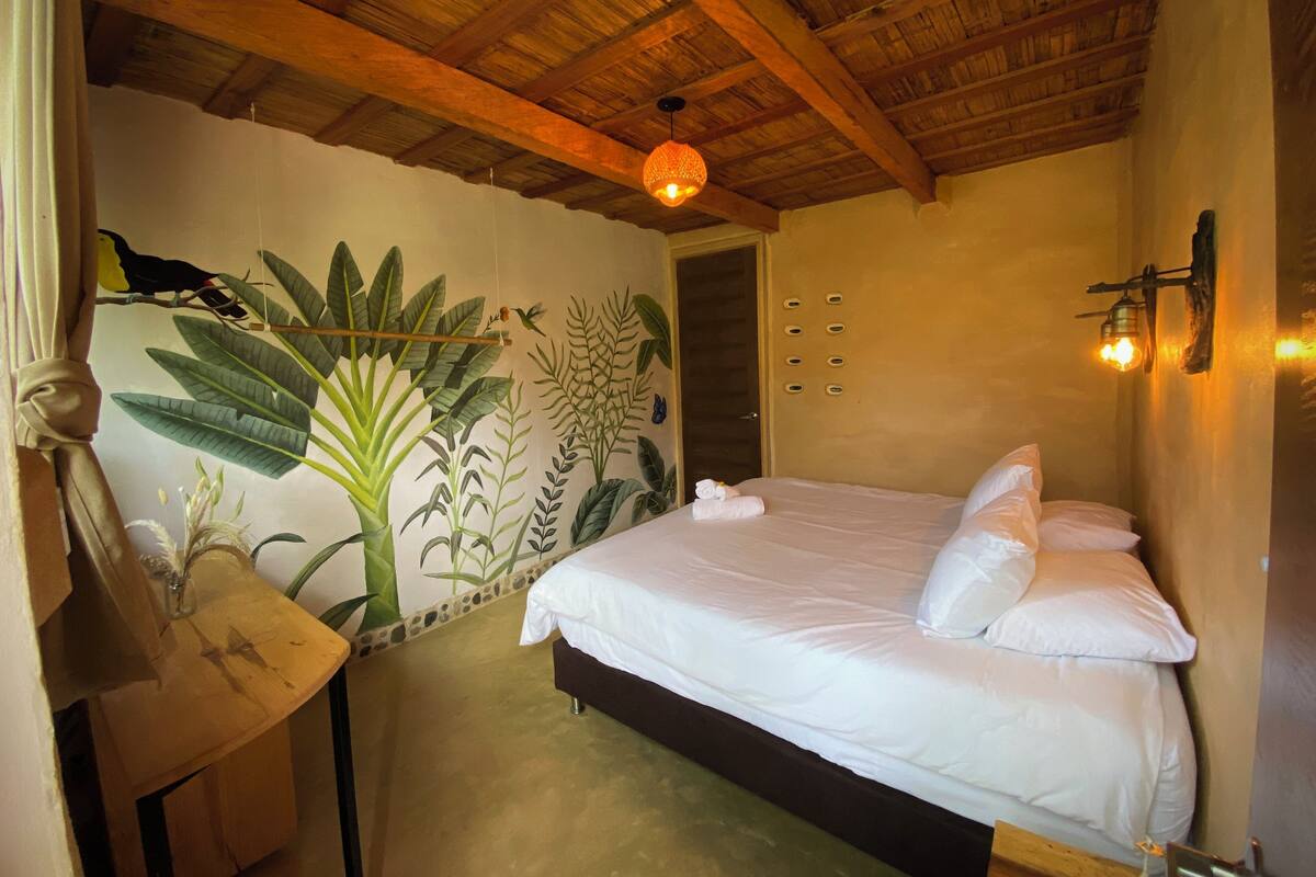 A bedroom with a bed, a wooden ceiling, a wall mural, and a small table.