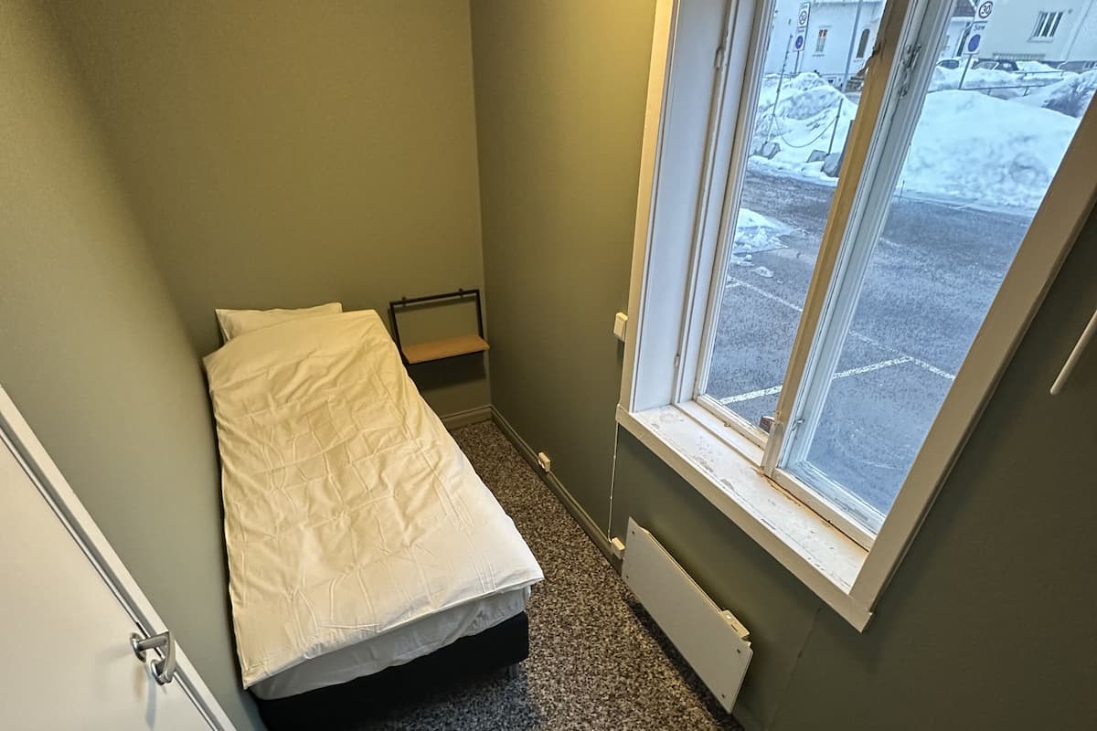 A small, compact room with a single bed, a desk, and a window providing a view of the outside.