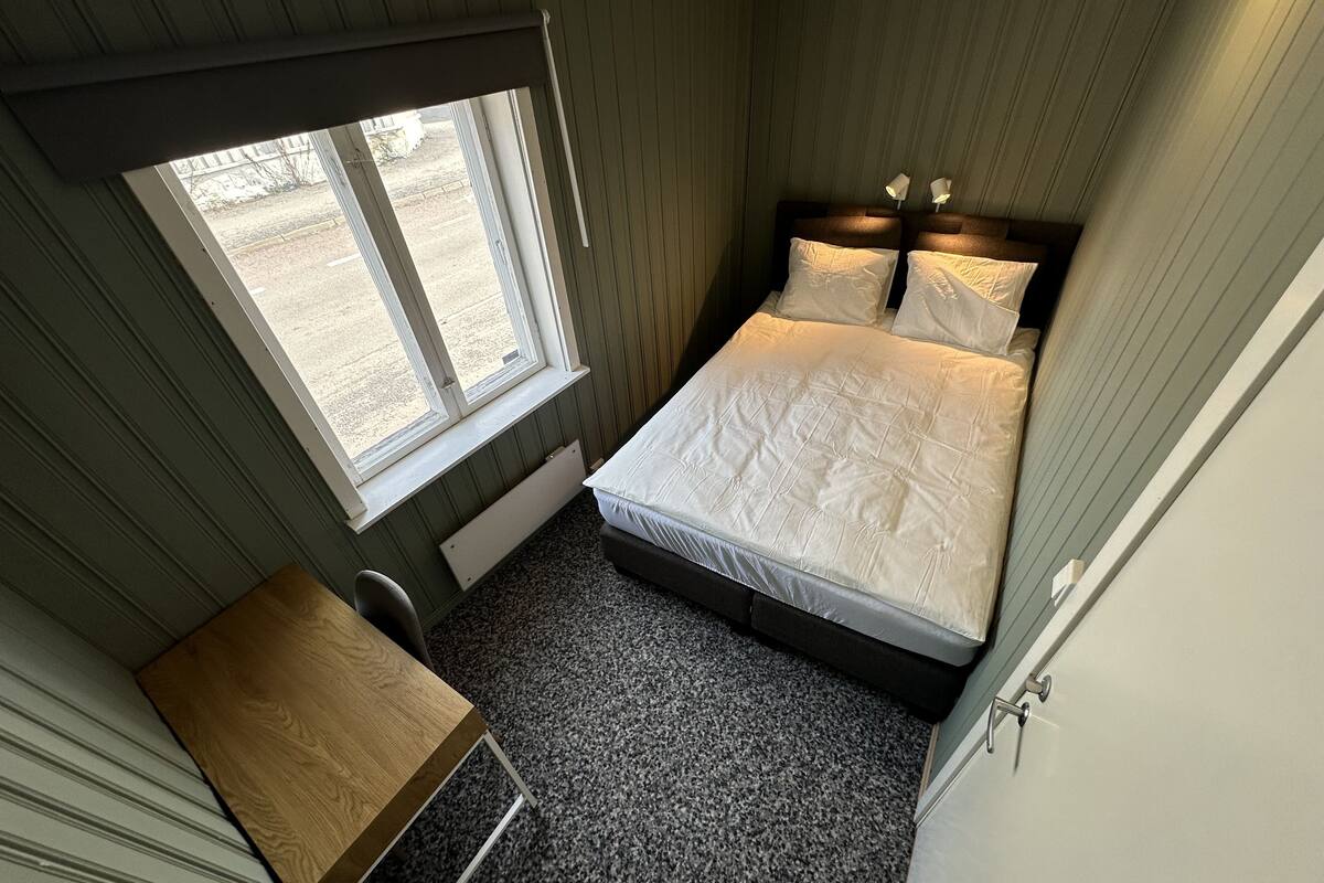 A compact bedroom with a single bed, a small desk, and a window providing a view of the outside.