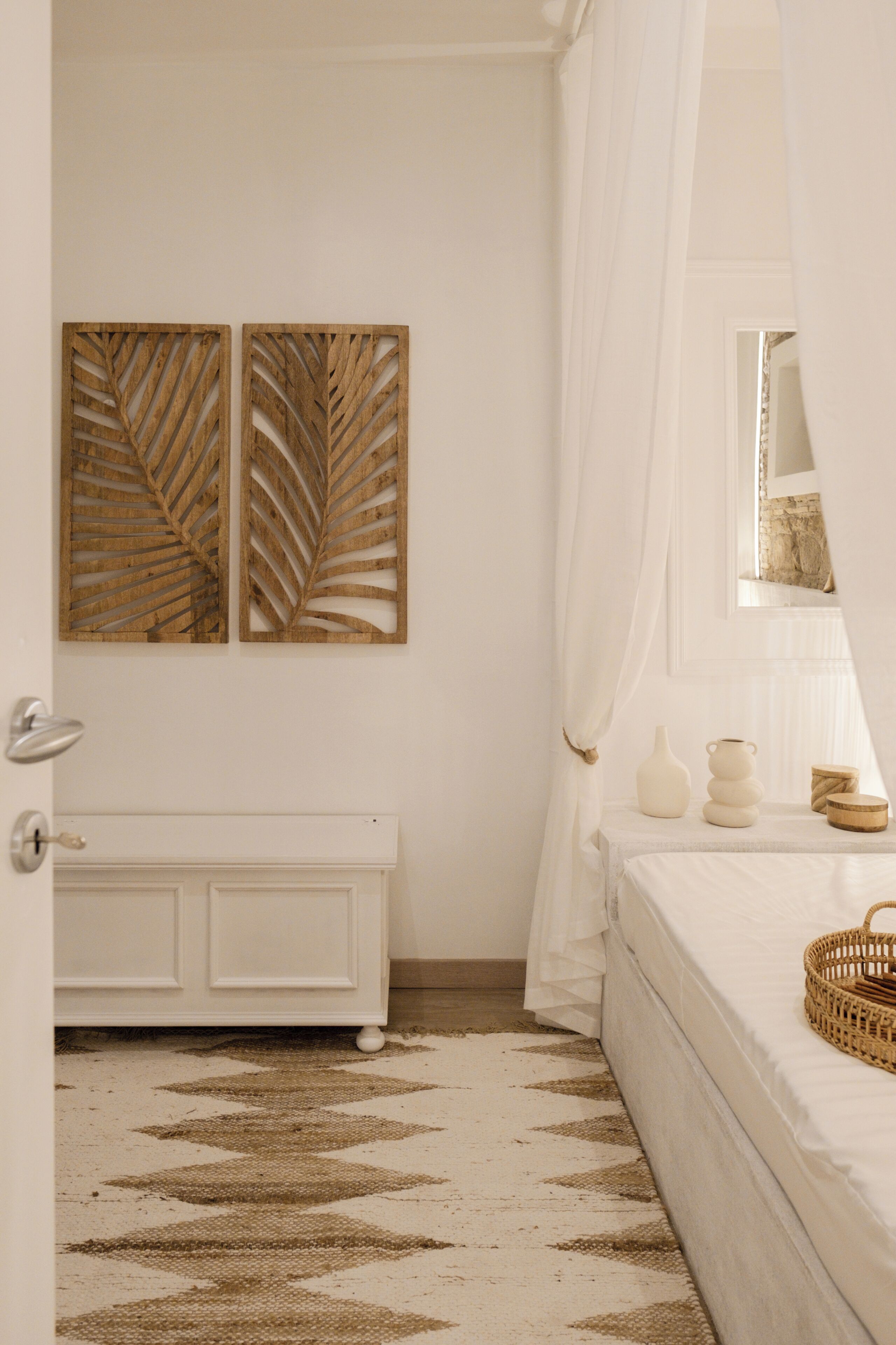 A bedroom with a white radiator, a patterned rug, a bed with a white bedspread, a bedside table with decorative items, and a window with sheer curtains.