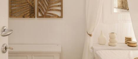 A bedroom with a white radiator, a patterned rug, a bed with a white bedspread, a bedside table with decorative items, and a window with sheer curtains.