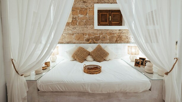A bedroom with a stone wall, a bed with white bedding, two brown pillows, a wicker basket, and a wooden cabinet.