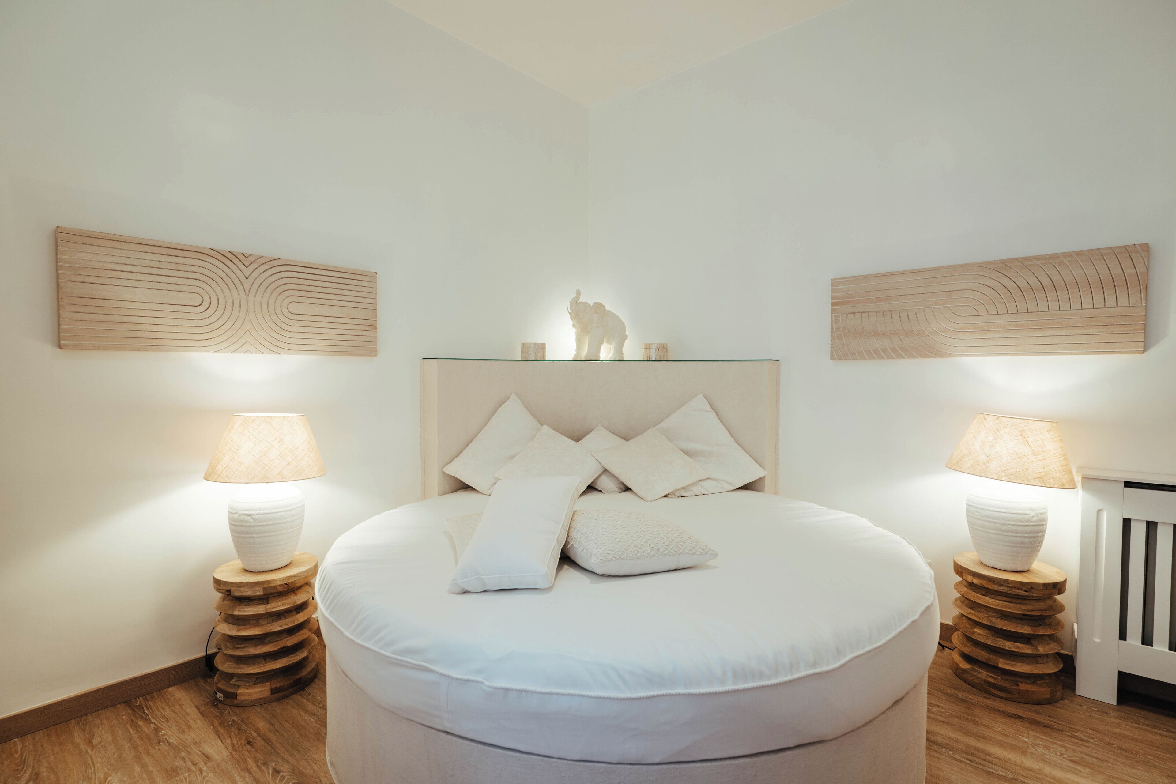 A bedroom with a round bed, two bedside tables with lamps, and wooden wall art above the bed.