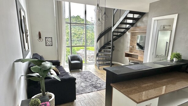 A modern kitchen with a central island, a staircase leading to an upper level, and a large window offering a view of greenery. - Saint Sebastian Flat 704Duplex no Centro (Jaragua do Sul)
