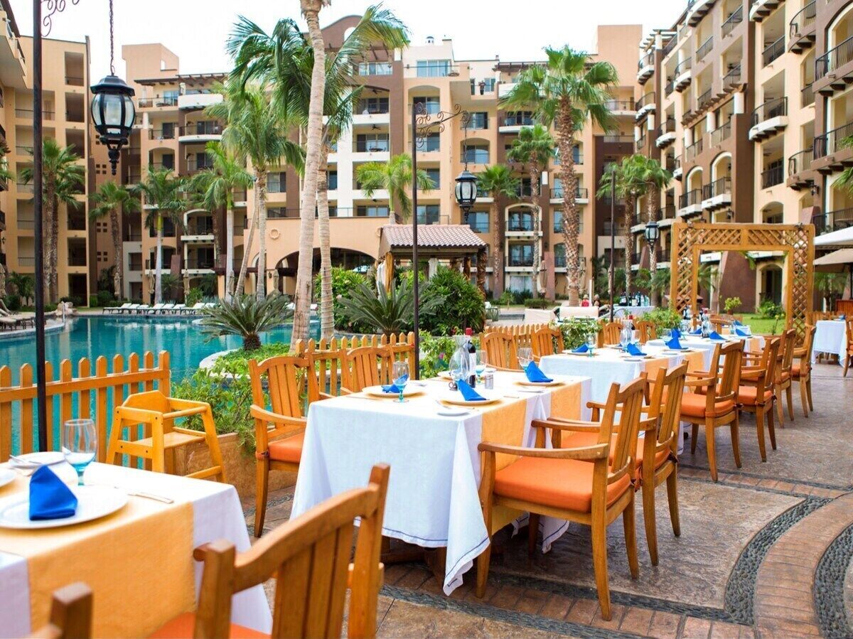 A poolside dining area with tables set for guests, surrounded by palm trees and a building with balconies.