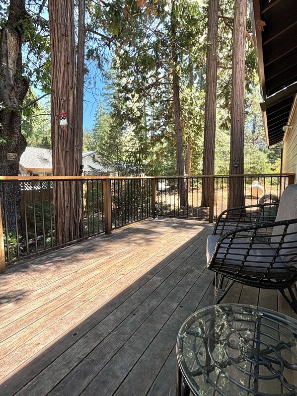 A wooden deck with a glass table and metal chair, surrounded by tall trees and a metal railing. - Cozy Mountain Getaway Near Sly Park Lake & Apple Hill + Game Garage (Pollock Pines)