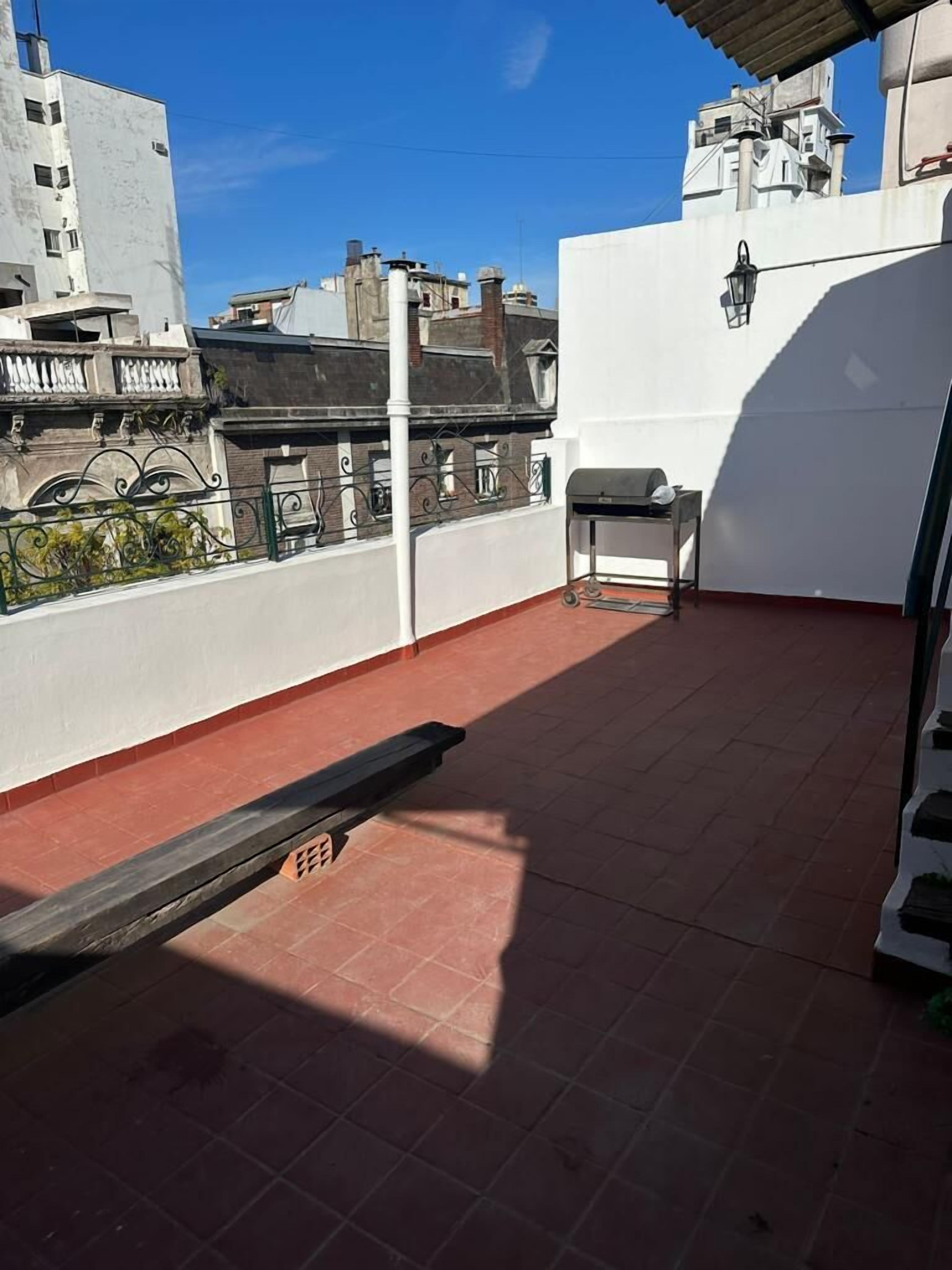 A rooftop terrace with a grill, a metal bench, and a view of surrounding buildings.
