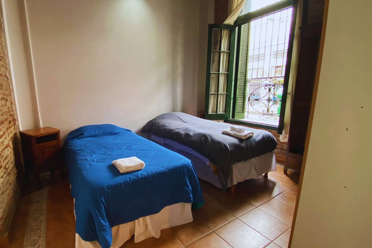 A room with two beds, a brick wall, a small wooden nightstand, and a window with a view of a bicycle and buildings.