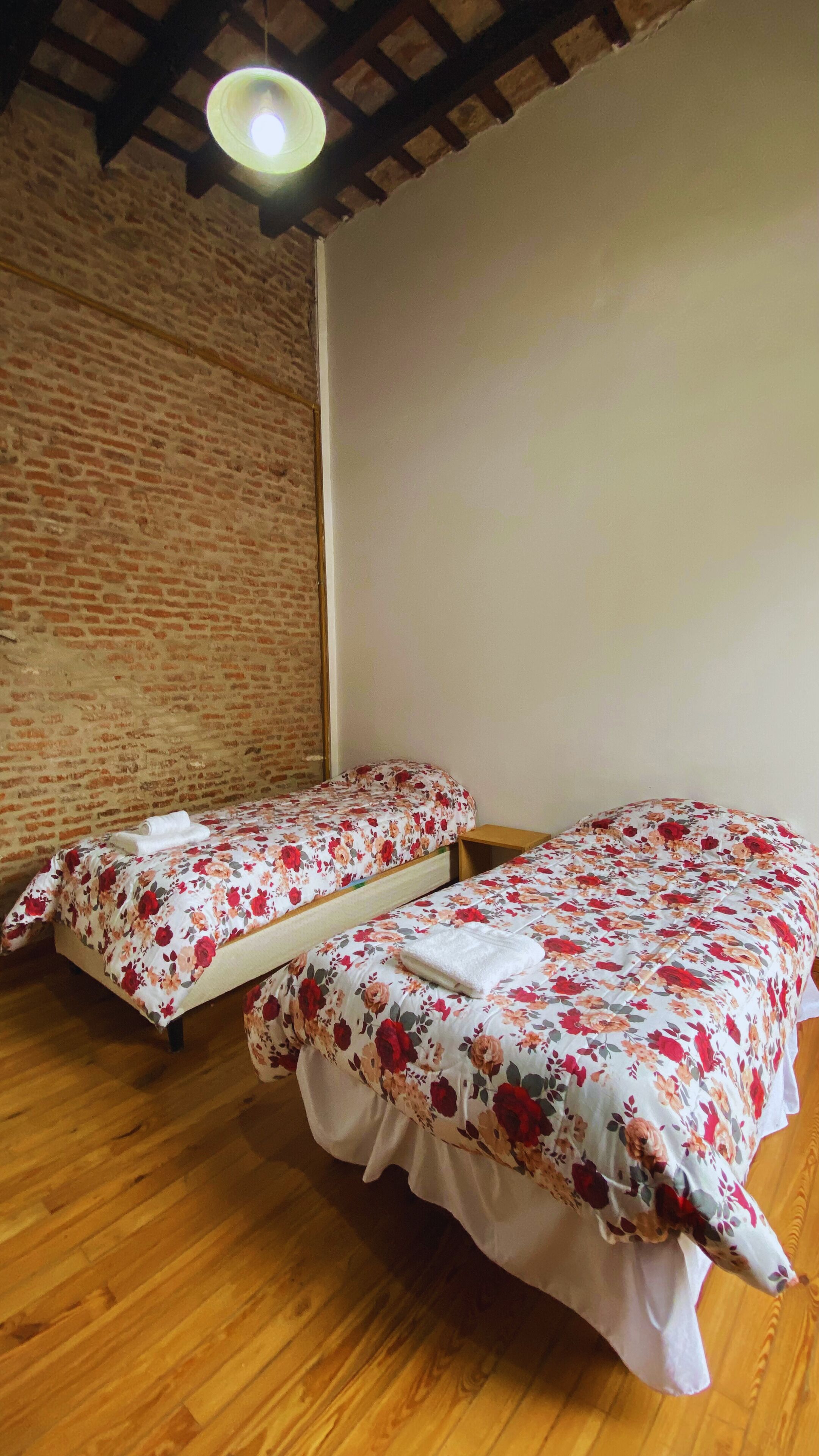 A room with two beds, wooden flooring, a brick wall, and a hanging light fixture.