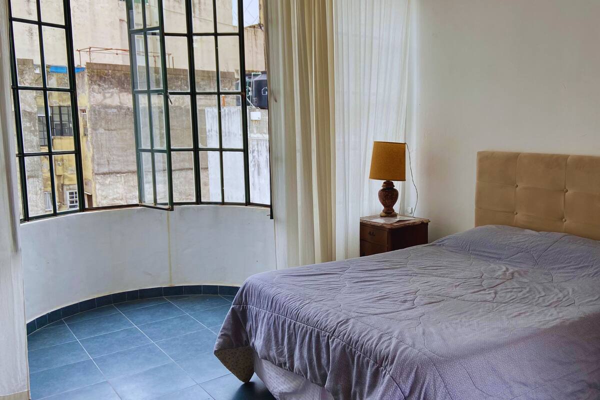 A room with a bed, a nightstand, a lamp, a window with curtains, and a view of buildings outside.