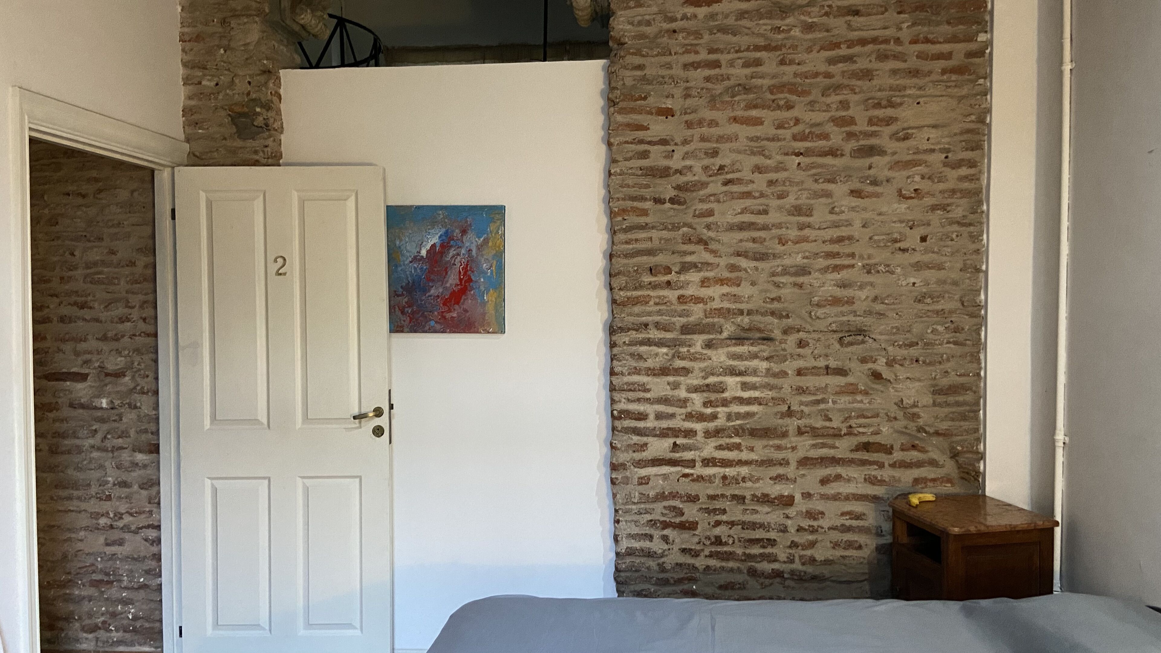 Una habitación con una puerta blanca marcada con el número 2, una pared de ladrillo, una pequeña mesa de madera y un cuadro colorido en la pared.