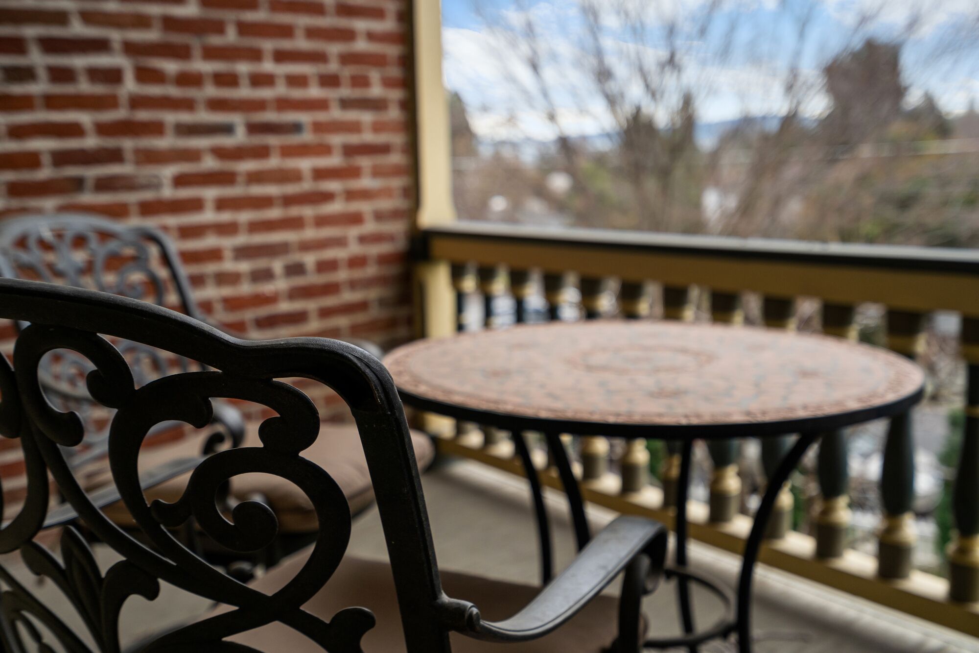 Een balkon met een ronde tafel en stoelen, een bakstenen muur en uitzicht op bomen en gebouwen.