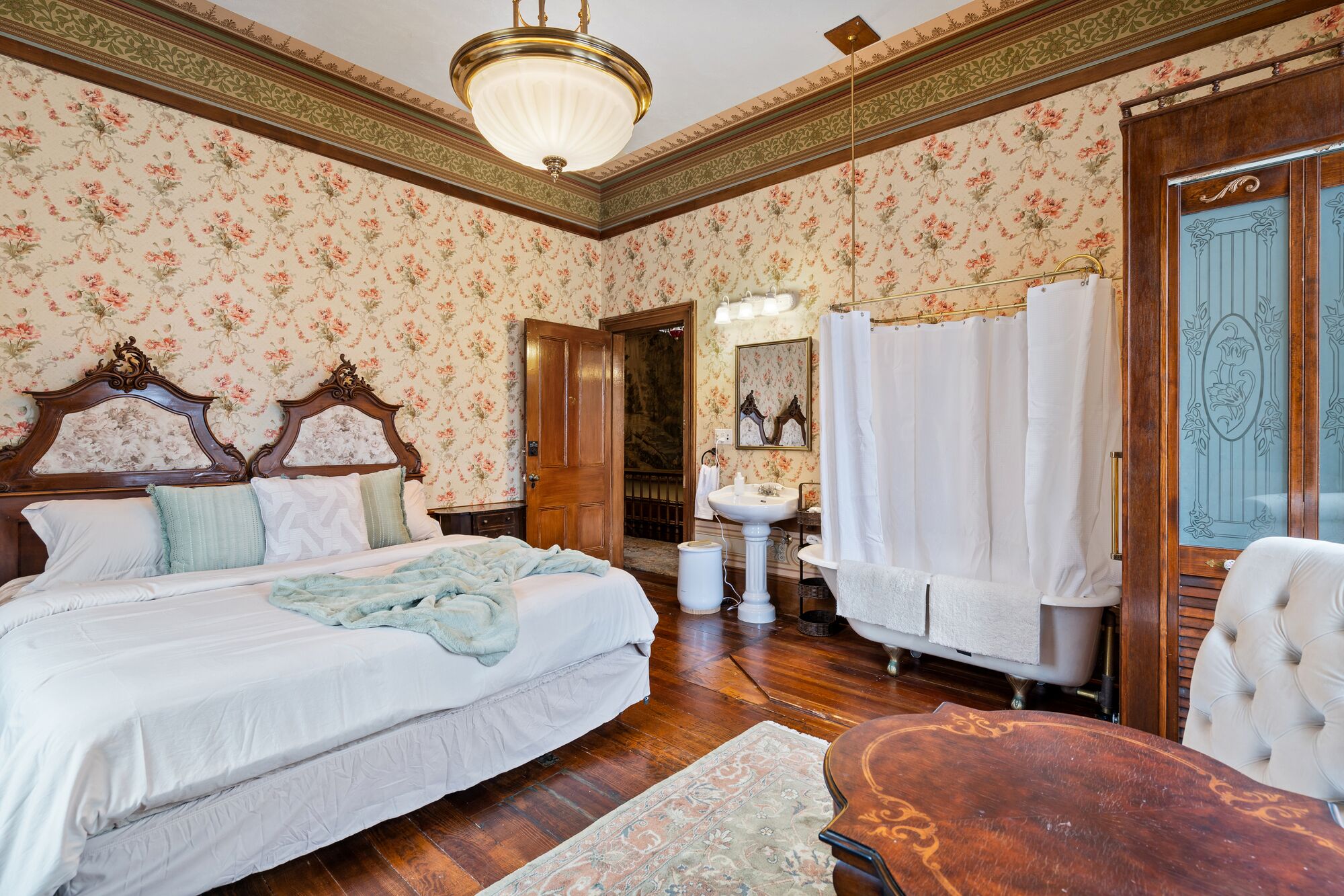 A bedroom with a large bed, a wooden headboard, a nightstand, a dresser, a vanity with a mirror, a bathtub, and a wooden chair.