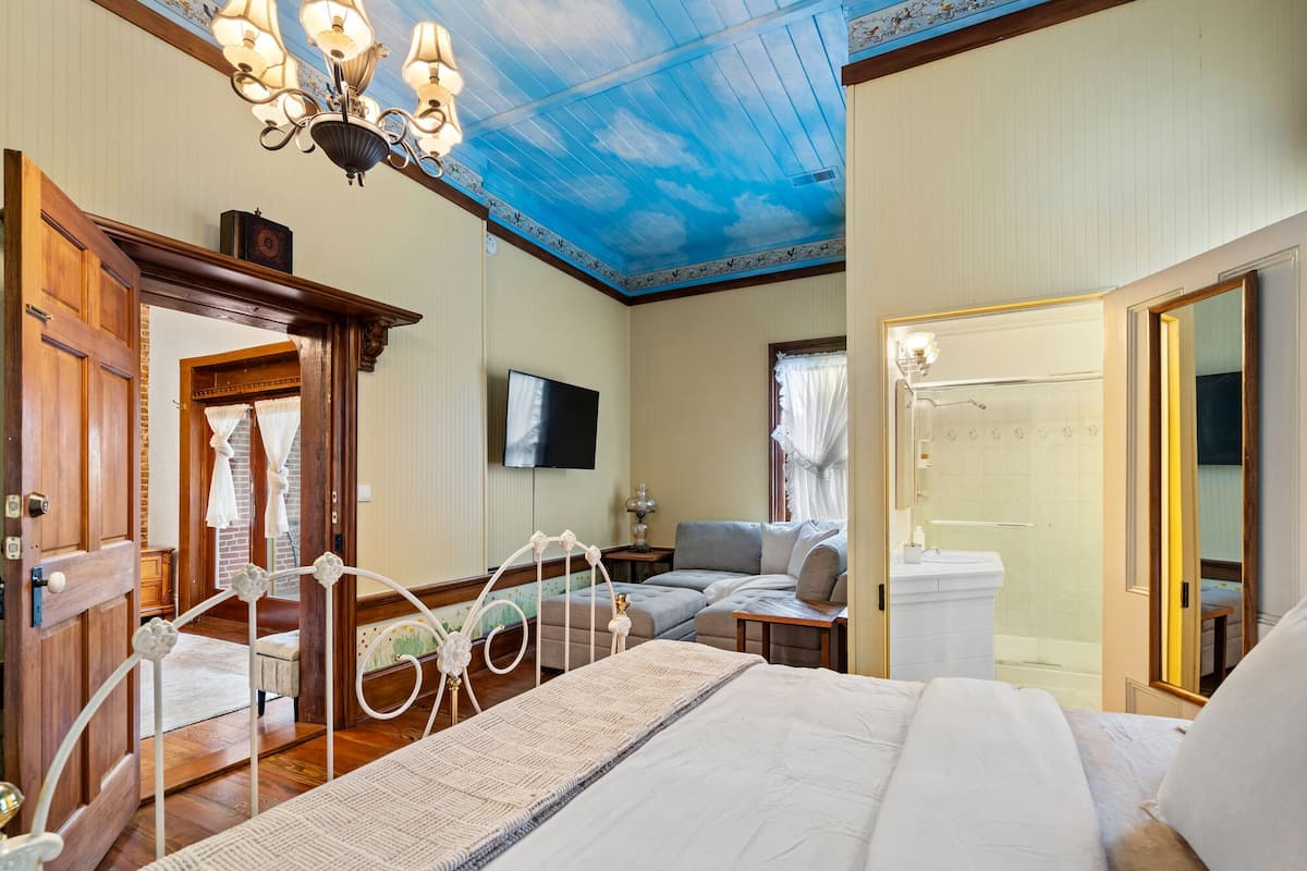 A hotel room with a bed, a sofa, a TV, a bathroom with a shower, and a ceiling painted to resemble the sky.