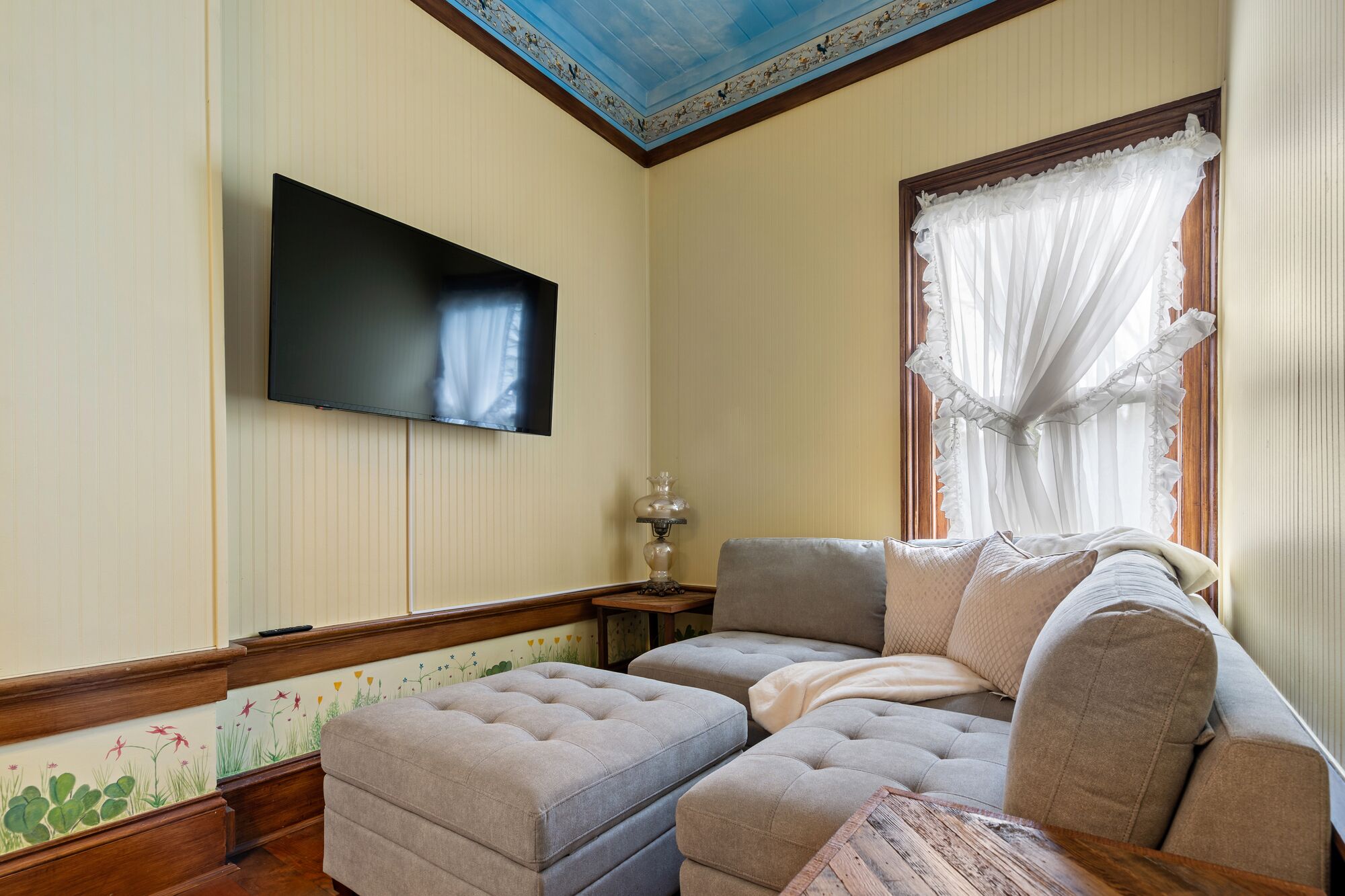 A living room with a sectional sofa, a flat-screen TV mounted on the wall, and a window with sheer curtains.